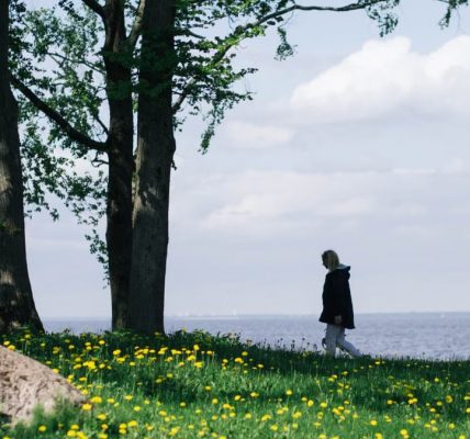 A woman walks along a grassy field by a serene lake under a clear spring sky.